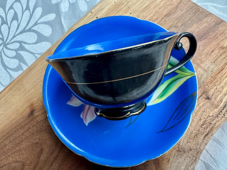 Puede incluir: Un juego de taza y platillo de t&eacute; azul y negro con un dise&ntilde;o floral. La taza de t&eacute; tiene un interior negro y un borde dorado. El platillo tiene un fondo azul con un dise&ntilde;o floral rosa y verde.