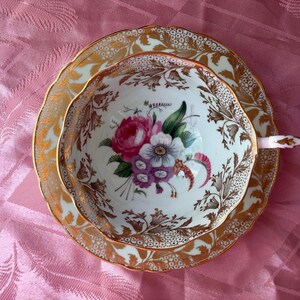 May include: A porcelain teacup and saucer set featuring a white base with a central floral bouquet of pink, purple, and white flowers. The rim and exterior are decorated with intricate gold leaf filigree and foliage patterns on a scalloped edge.