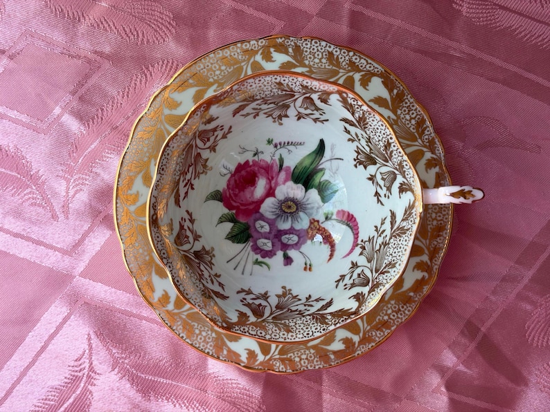 May include: A porcelain teacup and saucer set featuring a white base with a central floral bouquet of pink, purple, and white flowers. The rim and exterior are decorated with intricate gold leaf filigree and foliage patterns on a scalloped edge.