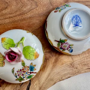 May include: A pair of porcelain lidded trinket boxes with a floral design. The lid of one box features a pink rose with green leaves and a butterfly with blue, white, and yellow wings. The bottom of the other box has a blue and white stamp that reads "Herend Hungary".