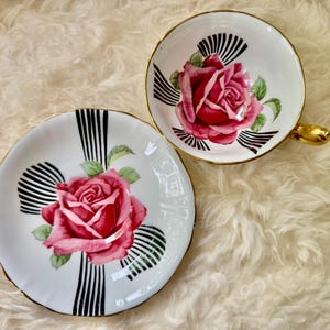 May include: A vintage teacup and saucer set. The white porcelain features a large pink rose in the center, accented by black and white striped designs. The cup has a gold handle and trim, and the saucer has a matching design.