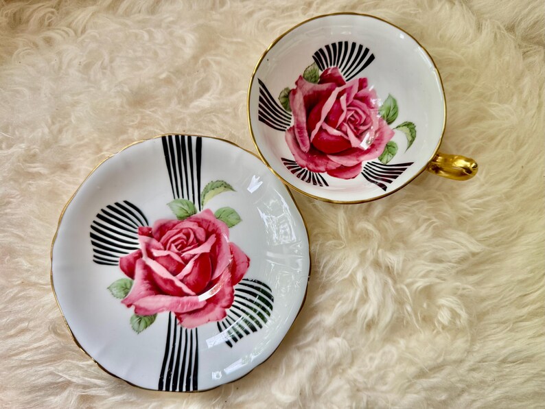 May include: A vintage teacup and saucer set. The white porcelain features a large pink rose in the center, accented by black and white striped designs. The cup has a gold handle and trim, and the saucer has a matching design.