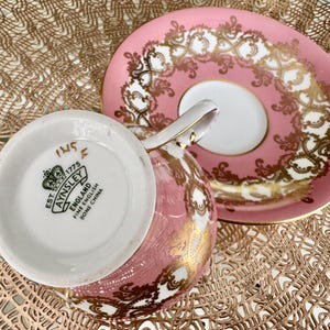 May include: Pink and gold Aynsley teacup and saucer set. The teacup is pink with gold filigree, and the saucer is pink with a white center and gold filigree. The bottom of the teacup has the Aynsley England Fine Bone China logo.