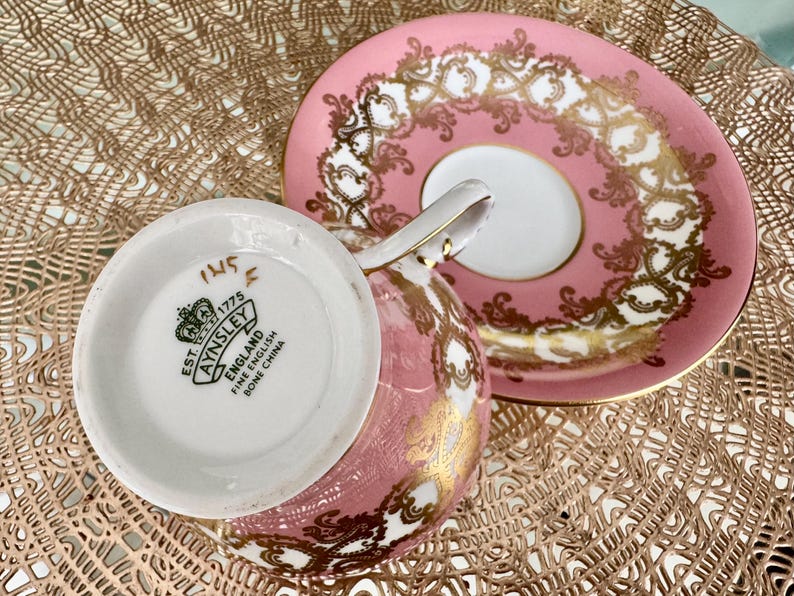 May include: Pink and gold Aynsley teacup and saucer set. The teacup is pink with gold filigree, and the saucer is pink with a white center and gold filigree. The bottom of the teacup has the Aynsley England Fine Bone China logo.