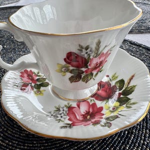 Puede incluir: Un juego de taza y platillo de t&eacute; blanco con un dise&ntilde;o floral con rosas rojas, rosas y amarillas. El juego tiene un borde dorado.