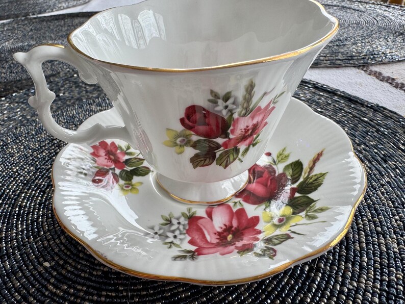 Puede incluir: Un juego de taza y platillo de t&eacute; blanco con un dise&ntilde;o floral con rosas rojas, rosas y amarillas. El juego tiene un borde dorado.