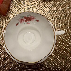 May include: A white teacup with a gold rim and a floral design featuring pink and red roses. The teacup is sitting on a gold-colored doily.