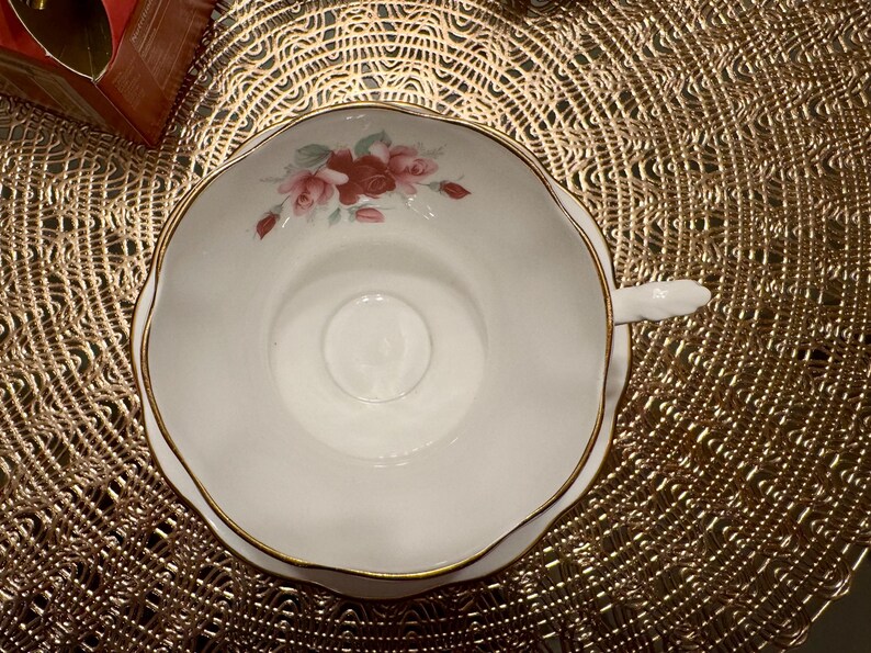 May include: A white teacup with a gold rim and a floral design featuring pink and red roses. The teacup is sitting on a gold-colored doily.