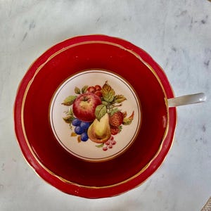 May include: A red teacup and saucer with a gold rim. The teacup features a white floral design with a pear, apple, and grapes.