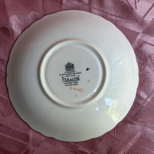 May include: A white bone china saucer with a scalloped edge. The underside features a black stamp with a royal crest, the text BY APPOINTMENT TO HER MAJESTY THE QUEEN, PARAGON, FINE BONE CHINA, ENGLAND REGD, and a handwritten orange code A3208/1.