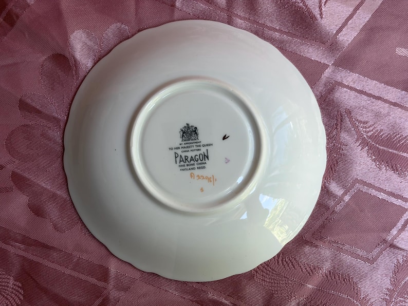 May include: A white bone china saucer with a scalloped edge. The underside features a black stamp with a royal crest, the text BY APPOINTMENT TO HER MAJESTY THE QUEEN, PARAGON, FINE BONE CHINA, ENGLAND REGD, and a handwritten orange code A3208/1.