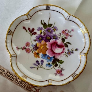 May include: A white, scalloped-edge dish with a floral design. The center features a bouquet of pink, purple, yellow, and blue flowers. The rim is trimmed with gold, and the dish has a decorative shape.