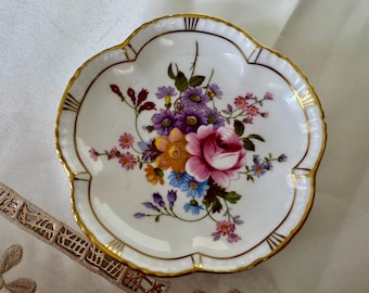 May include: A white, scalloped-edge dish with a floral design. The center features a bouquet of pink, purple, yellow, and blue flowers. The rim is trimmed with gold, and the dish has a decorative shape.