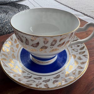 May include: A white teacup and saucer with a blue and gold floral pattern. The teacup has a gold rim and a gold floral design on the outside. The saucer has a blue center with a gold rim and a gold floral design on the outside.