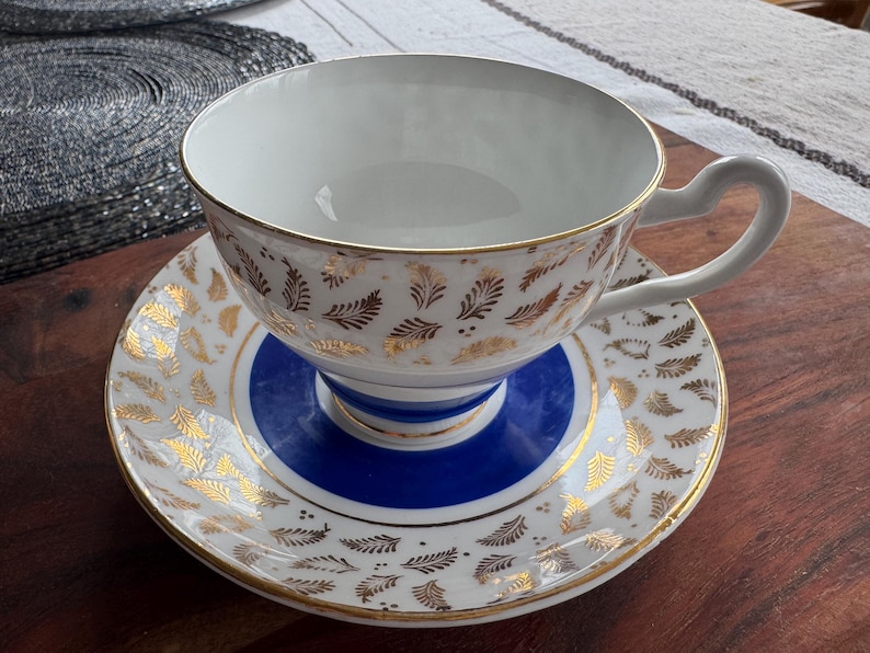 May include: A white teacup and saucer with a blue and gold floral pattern. The teacup has a gold rim and a gold floral design on the outside. The saucer has a blue center with a gold rim and a gold floral design on the outside.