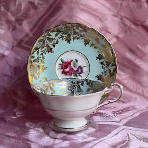 May include: A porcelain teacup and saucer set featuring a pale mint green background with intricate gold filigree floral patterns. The center of the saucer displays a small bouquet of pink, white, and purple flowers. The teacup has a gold-trimmed rim.