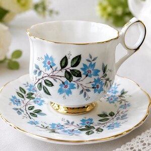 May include: A white porcelain teacup and saucer set featuring a delicate pattern of light blue flowers and green leaves. The teacup has a scalloped rim, a gold-trimmed handle, and a gold base, while the matching saucer also has a gold-trimmed edge.