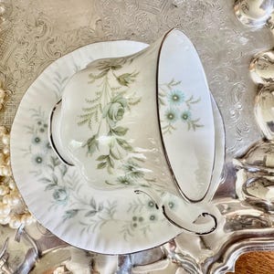 May include: A white teacup and saucer set with a floral design in shades of green and blue, accented with gold trim. The teacup is resting on a matching saucer, placed on a silver tray. A strand of pearls is visible.