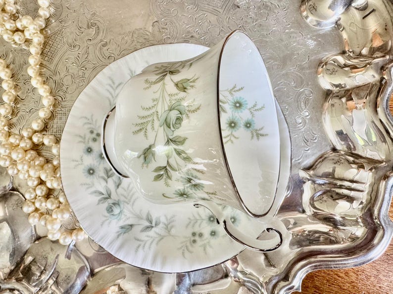May include: A white teacup and saucer set with a floral design in shades of green and blue, accented with gold trim. The teacup is resting on a matching saucer, placed on a silver tray. A strand of pearls is visible.