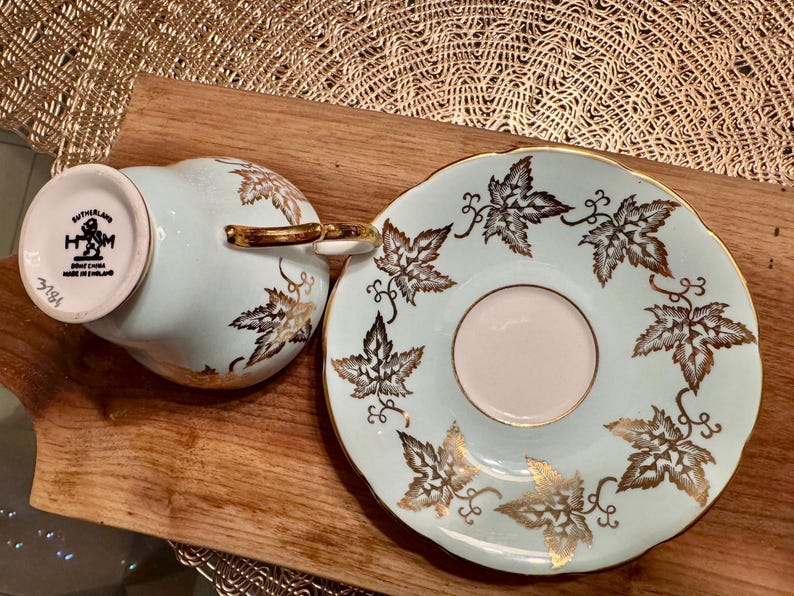 May include: A light blue teacup and saucer set with a gold leaf design. The teacup has a white underside with the text "Sutherland Made China Made in England 3181".