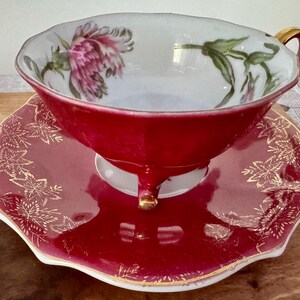 May include: A vintage teacup and saucer set. The teacup is a deep red with a gold handle and foot. The interior of the cup and the saucer are white with a floral design. The saucer is red with a gold leaf pattern around the edge. A classic tea set.