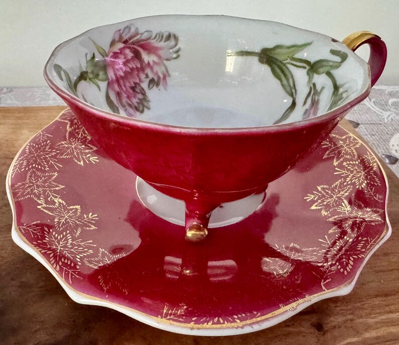 May include: A vintage teacup and saucer set. The teacup is a deep red with a gold handle and foot. The interior of the cup and the saucer are white with a floral design. The saucer is red with a gold leaf pattern around the edge. A classic tea set.