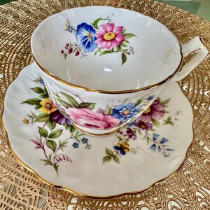 May include: A white teacup and saucer with a floral pattern in shades of pink, blue, yellow, and green. The cup and saucer have a gold trim.
