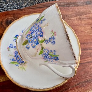 May include: A white teacup and saucer with a gold rim and a floral pattern of blue forget-me-nots.