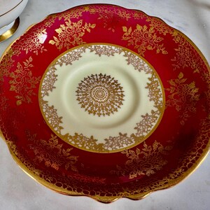 May include: A red and gold vintage teacup saucer and a smaller cream-colored teacup saucer with a gold floral design. The edges of the saucers are scalloped and trimmed with gold. The teacup saucer has a gold floral design.