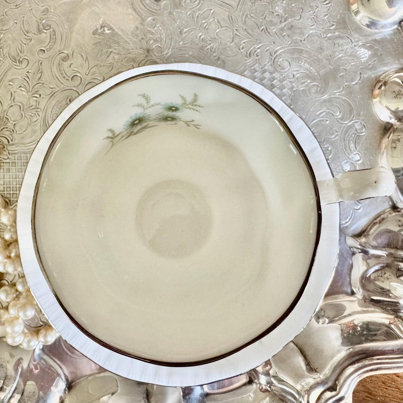 May include: A cream-colored teacup with a delicate floral design in shades of green and blue. The cup is set within a silver-colored holder, creating a vintage aesthetic. The teacup is placed on a decorative silver tray.