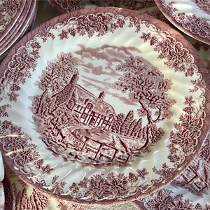 Pu&ograve; includere: Una collezione di piatti in ceramica bianchi e bordeaux. Ogni piatto presenta una scena paesaggistica dettagliata con una casa, alberi e una recinzione. I piatti hanno un bordo floreale decorativo e sono impilati.
