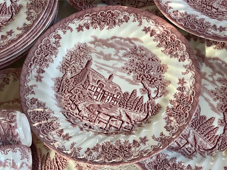 Pu&ograve; includere: Una collezione di piatti in ceramica bianchi e bordeaux. Ogni piatto presenta una scena paesaggistica dettagliata con una casa, alberi e una recinzione. I piatti hanno un bordo floreale decorativo e sono impilati.