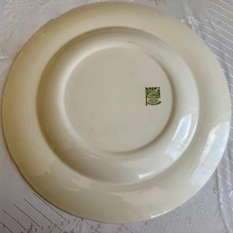 May include: Off-white ceramic plate with a raised rim and a central recessed area. The underside features a green stamp with a Union Jack and text. The plate is likely vintage or antique, showcasing a classic design.