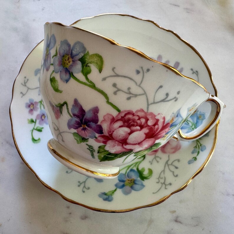 Pu&ograve; includere: Tazza da t&egrave; e piattino bianchi con decorazioni floreali. La tazza e il piattino hanno un bordo smerlato e rifiniture dorate. Il motivo floreale include fiori rosa, viola e blu con foglie verdi.