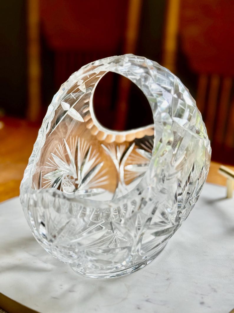 May include: A clear, cut-glass basket with a handle. The basket features intricate starburst and geometric patterns. The handle has a large opening. The basket is likely used for decorative purposes or to hold small items.