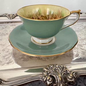 May include: A vintage teacup and saucer set. The cup and saucer are a muted green with gold trim. The inside of the cup is a light yellow with a floral design. The set is placed on a silver tray.