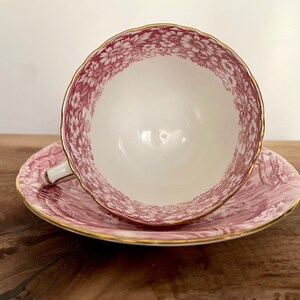 Pu&ograve; includere: Un set vintage di tazza e piattino da t&egrave;. La tazza e il piattino sono bianchi con un motivo floreale rosa e finiture dorate. La tazza &egrave; appoggiata sul piattino. Il set &egrave; su una superficie di legno.