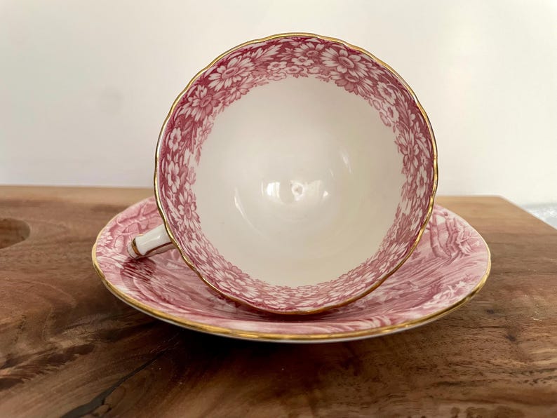 Pu&ograve; includere: Un set vintage di tazza e piattino da t&egrave;. La tazza e il piattino sono bianchi con un motivo floreale rosa e finiture dorate. La tazza &egrave; appoggiata sul piattino. Il set &egrave; su una superficie di legno.
