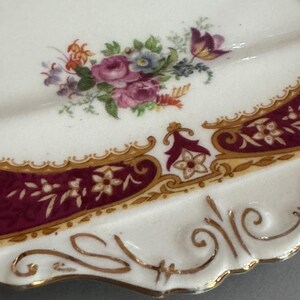 May include: A white porcelain plate with a burgundy and gold border. The plate features a floral design with pink, red, and blue flowers.