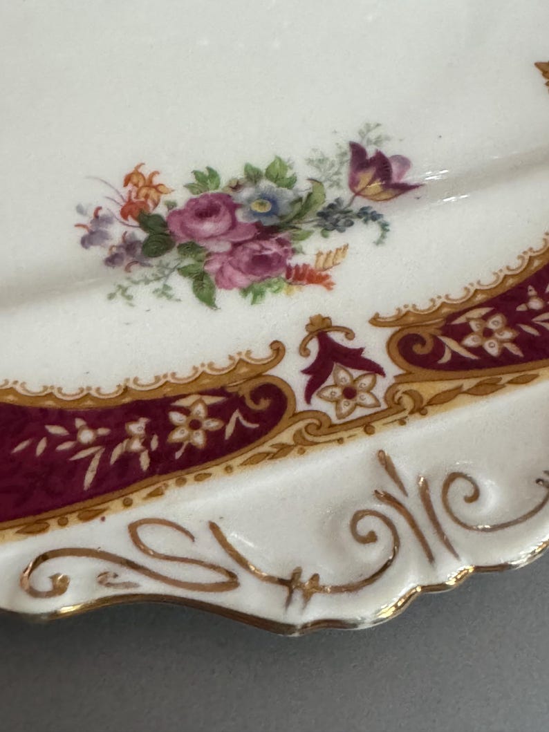 May include: A white porcelain plate with a burgundy and gold border. The plate features a floral design with pink, red, and blue flowers.