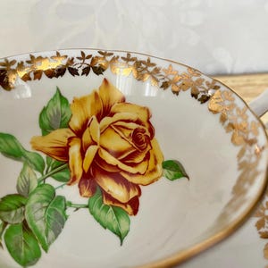 Puede incluir: Taza de t&eacute; blanca con un dise&ntilde;o de rosa amarilla. La taza tiene un borde y un asa dorados, y la rosa est&aacute; rodeada de hojas verdes. La taza de t&eacute; es probablemente parte de un juego de t&eacute; vintage.