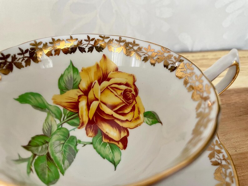 Puede incluir: Taza de t&eacute; blanca con un dise&ntilde;o de rosa amarilla. La taza tiene un borde y un asa dorados, y la rosa est&aacute; rodeada de hojas verdes. La taza de t&eacute; es probablemente parte de un juego de t&eacute; vintage.