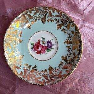May include: A round porcelain saucer with a scalloped edge, featuring a pale mint green background. The center displays a small floral bouquet of red, white, and purple flowers. The rim is decorated with intricate gold filigree and floral patterns.