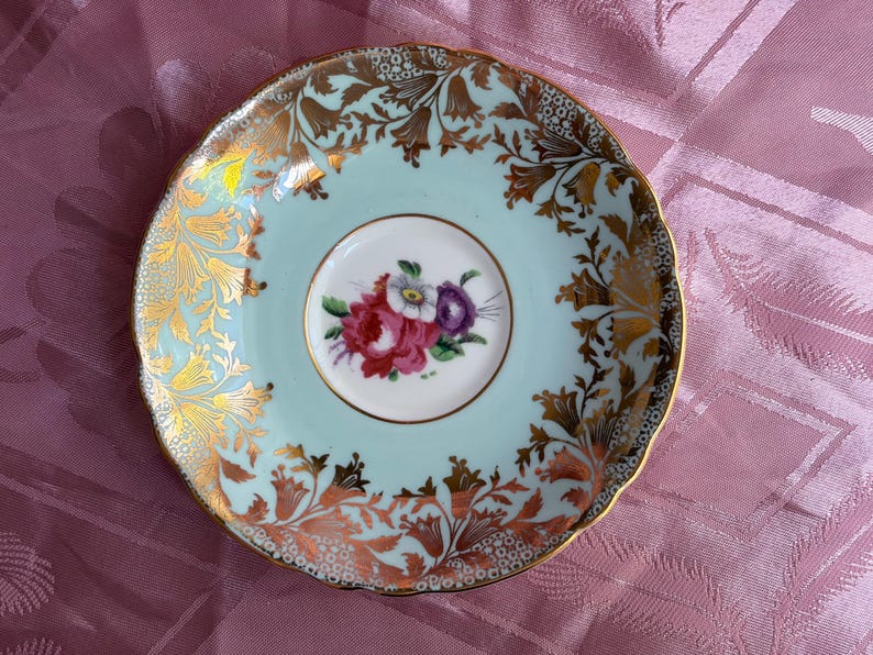 May include: A round porcelain saucer with a scalloped edge, featuring a pale mint green background. The center displays a small floral bouquet of red, white, and purple flowers. The rim is decorated with intricate gold filigree and floral patterns.
