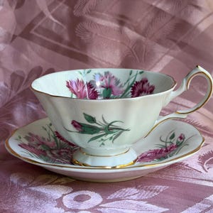 May include: A white porcelain teacup and saucer set featuring a scalloped edge and gold trim. The exterior of the cup and the surface of the saucer are decorated with a botanical pattern of pink carnations and green stems on a cream background.