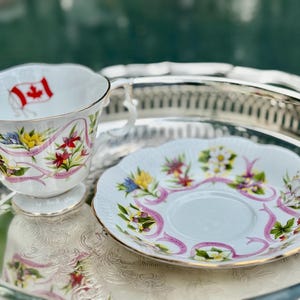 May include: A white teacup and saucer set with floral and ribbon designs. The teacup features a Canadian flag and gold trim. The saucer and cup are decorated with colorful flowers and pink ribbons. The set is placed on a silver tray.