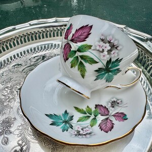 May include: A white teacup and saucer set with a floral design featuring pink, green, and white flowers and leaves. The teacup and saucer have gold trim and are placed on a silver serving tray.