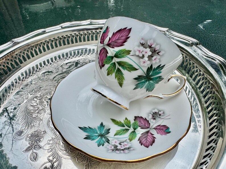 May include: A white teacup and saucer set with a floral design featuring pink, green, and white flowers and leaves. The teacup and saucer have gold trim and are placed on a silver serving tray.