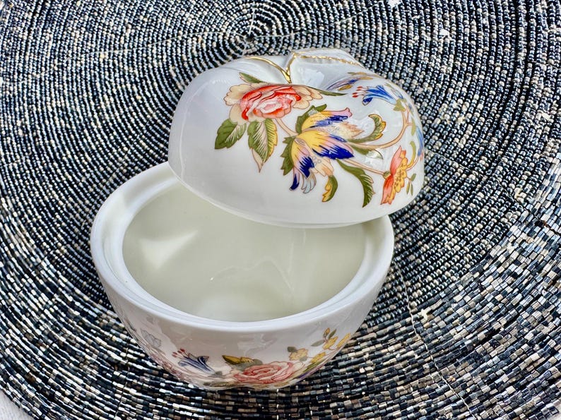 K&ouml;nnte beinhalten: Eine wei&szlig;e Porzellan-Schmuckdose in Apfelform mit floralem Design. Der Deckel ist ge&ouml;ffnet und das Innere der Dose ist sichtbar.