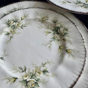 May include: A set of white ceramic plates with a delicate floral design featuring white roses and green leaves. The plates have a silver rim.